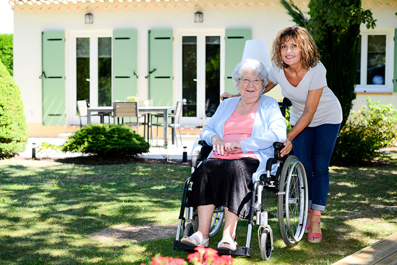 Polnische Betreuerin unterstützt ältere Frau beim Rollstuhlfahren in ihrem Garten.